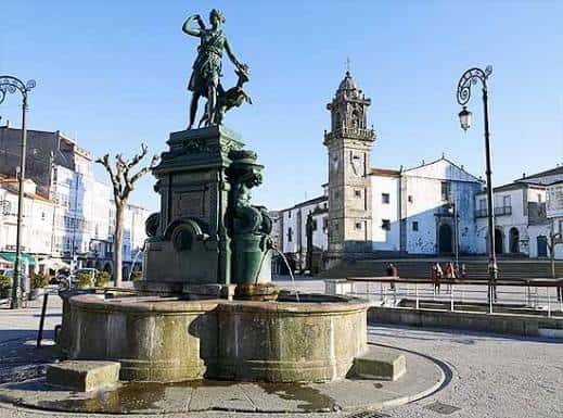 Fuente monumental en el centro de la plaza con estatua y torre histórica al fondo