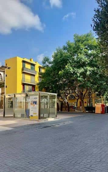Plaza adoquinada con árboles frondosos y parada de autobús bajo un cielo azul despejado