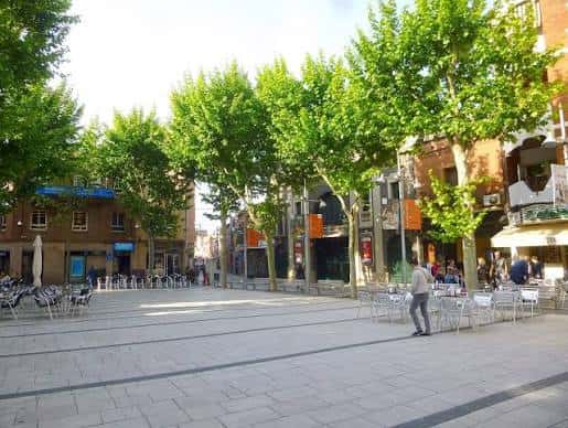 Vista diurna de la Plaça de la Vila con árboles frondosos y mesas de terraza