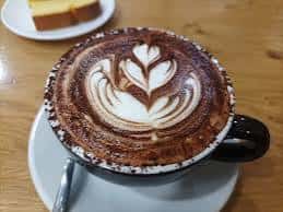 Taza de café con diseño de latte art y trozo de pastel al fondo
