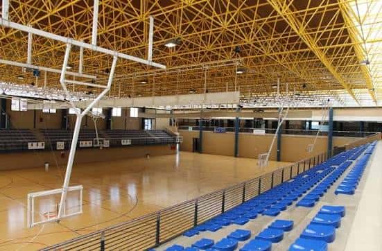 Interior del pabellón mostrando la cancha de baloncesto y gradas azules bajo una estructura metálica amarilla