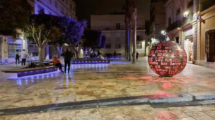 Plaza iluminada por la noche con escultura esférica y bancos modernos