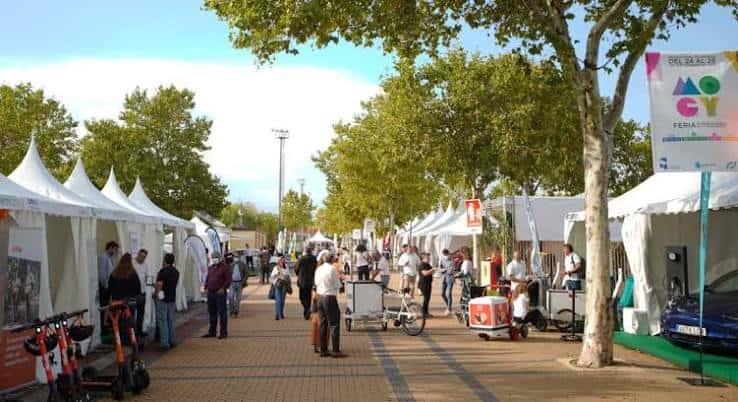 Pasillo central del recinto con carpas blancas, árboles y visitantes caminando