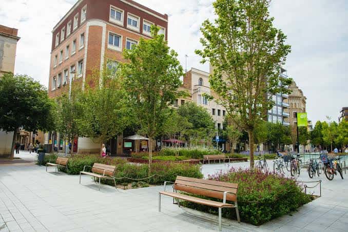 Vista diurna del Passeig de la Plaça Major con bancos y zonas ajardinadas