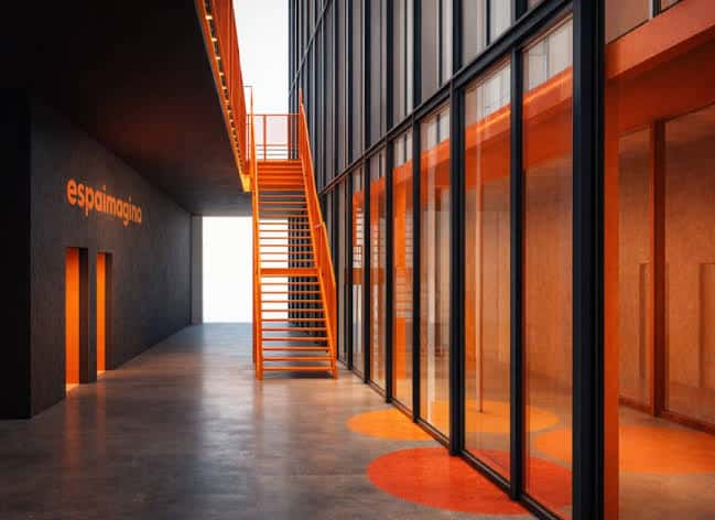 Interior moderno del Espai Imagina con escalera naranja y paredes de cristal que reflejan luz ambiental
