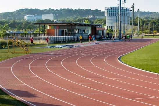 Pista de atletismo en primer plano con atletas entrenando y edificio moderno al fondo