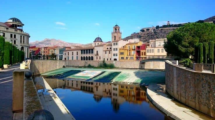 Vista diurna del Puente Nuevo en Orihuela con reflejos en el río Segura y la ciudad al fondo