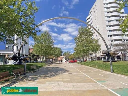 Paseo peatonal con escultura metálica arqueada y zonas verdes en la Plaça de la Llibertat