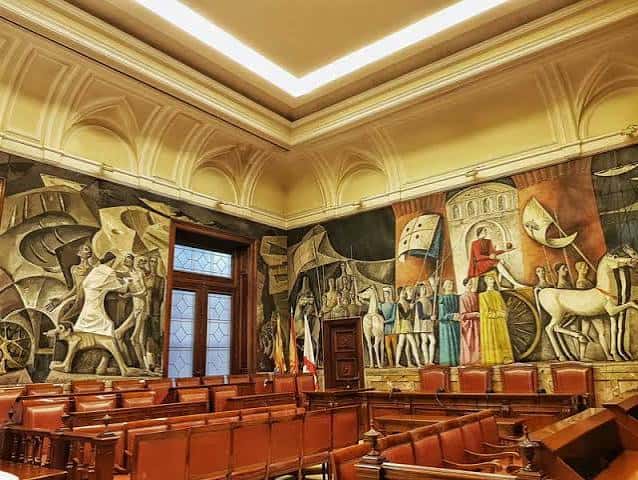 Interior del antiguo salón de plenos con murales históricos y butacas de madera