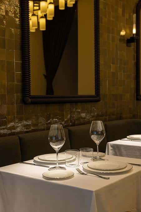Mesa servida para cena con vajilla blanca y copas de vino frente a pared de azulejos