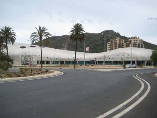 Fachada exterior del pabellón deportivo rodeado de palmeras y montañas al fondo