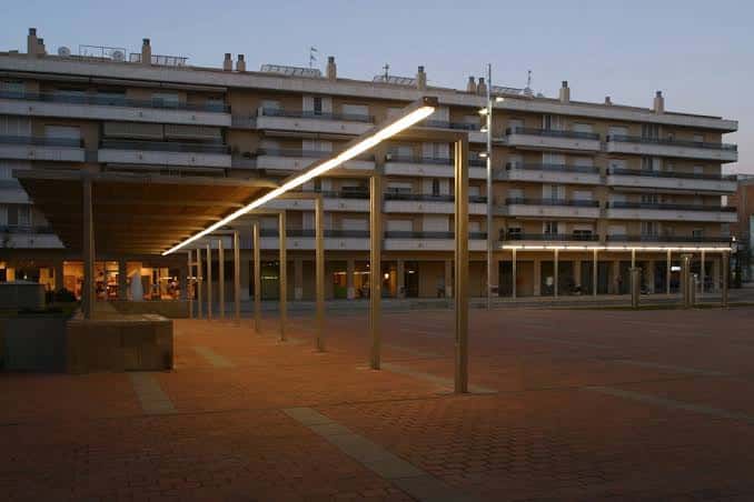 Pasarela iluminada en la plaza al atardecer con edificios residenciales al fondo