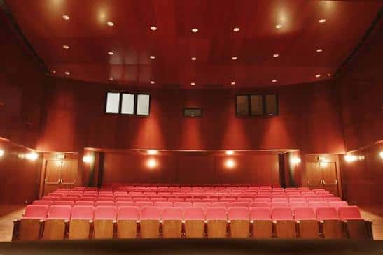 Sala de espectáculos vacía con butacas rojas y escenario iluminado