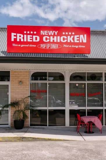 Vista frontal del local de Newy Fried Chicken con letrero rojo y mesas exteriores