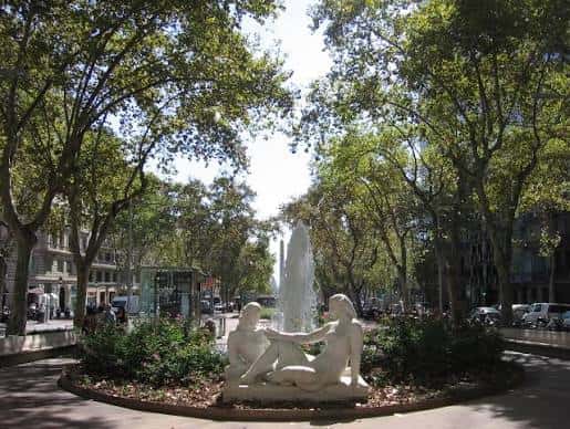 Vista del jardín con fuente central y escultura de figuras humanas bajo árboles frondosos
