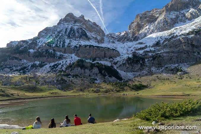 Grupo de personas descansando junto al ibón con montañas nevadas al fondo