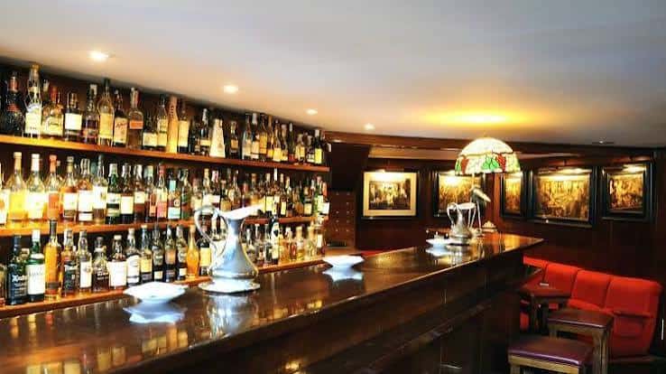 Interior del bar con estanterías llenas de botellas, barra de madera y sillones rojos