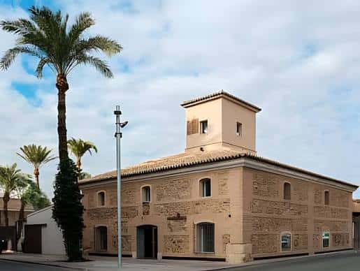 Fachada principal de la Casa del Belén en Murcia con palmeras y cielo despejado
