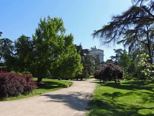 Camino entre vegetación variada con edificio blanco al fondo bajo cielo azul