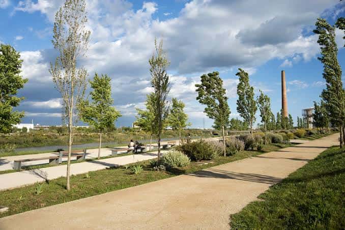 Paseo arbolado junto al río con bancos y la chimenea de fondo bajo cielo nublado