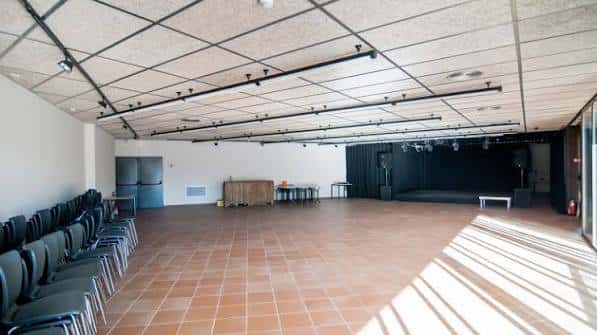 Sala polivalente interior con escenario, sillas apiladas y suelo de baldosas terracota