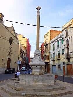 Primer plano de la columna conmemorativa y fuente en el centro de la plaza, rodeada de arquitectura tradicional