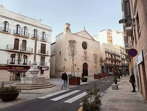 Plaza durante el día con peatones caminando junto a la iglesia y la cruz monumental, mostrando ambiente urbano tranquilo
