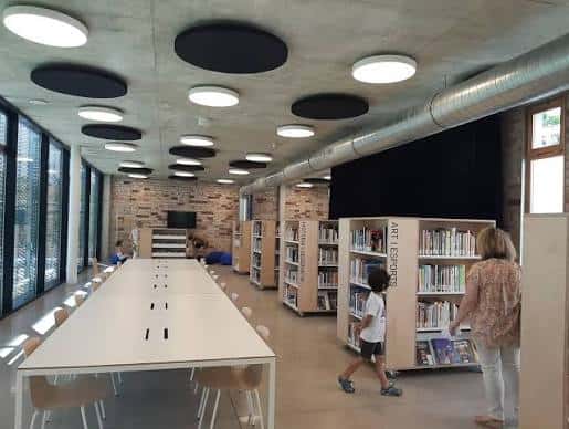 Interior de la biblioteca con mesas largas, estanterías y personas usando el espacio