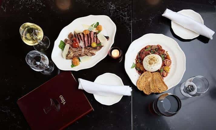 Platos gourmet de carne y ensalada servidos en mesa oscura con vino y menú