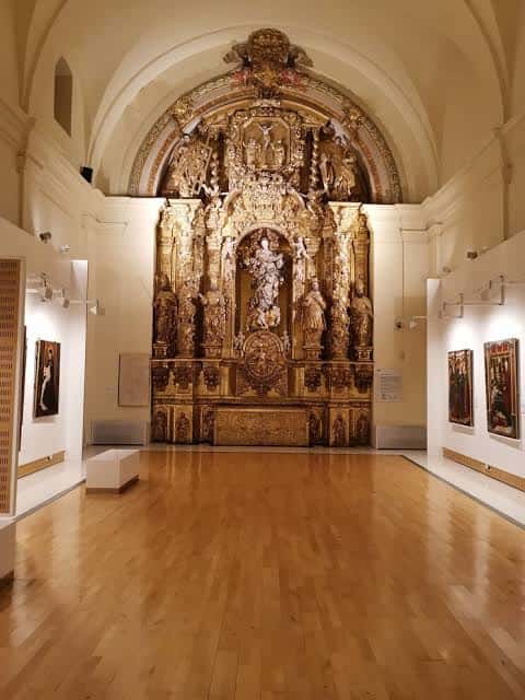 Sala de exposiciones con retablo dorado y obras de arte en las paredes