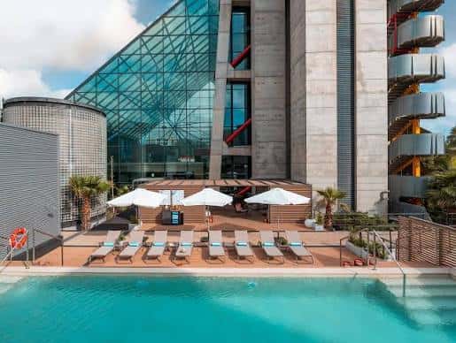 Piscina exterior del hotel con tumbonas, sombrillas y vista a la arquitectura de cristal del edificio