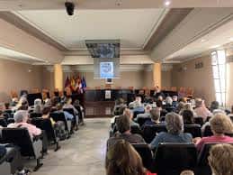 Sesión en el Salón de Plenos con público asistente y pantalla proyectando contenido
