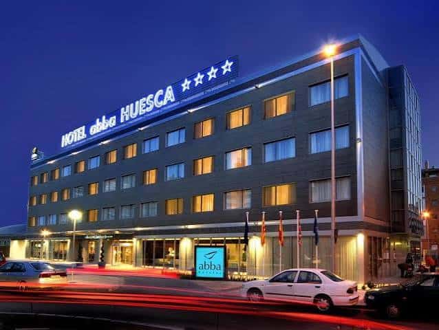 Fachada del hotel iluminada por la noche con luces cálidas en las ventanas y tráfico borroso en primer plano