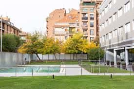 Área de piscina al aire libre rodeada de césped y árboles con edificios urbanos al fondo