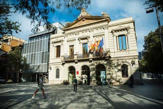 Fachada principal del edificio de la Plaça de Comas con banderas y árboles en primer plano