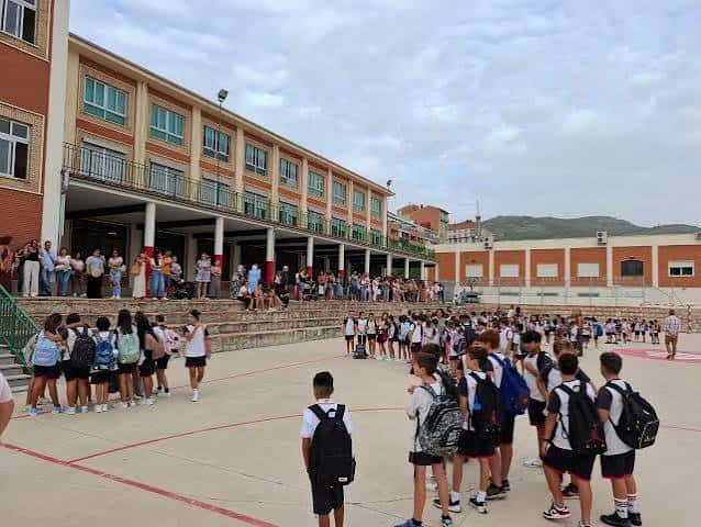 Patio exterior del colegio con alumnos en formación y gradas bajo cielo nublado