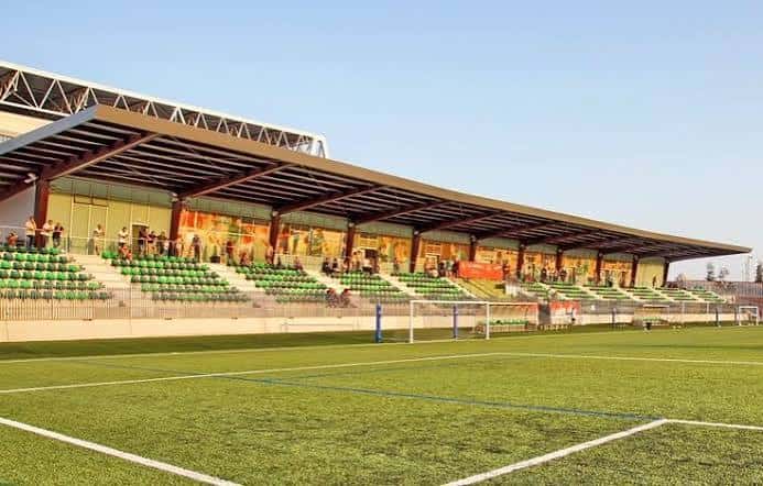 Gradas laterales del estadio con espectadores y murales decorativos bajo techo metálico