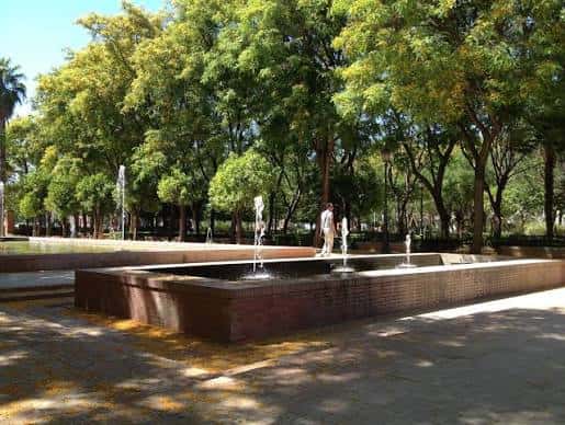 Fuente de ladrillo con chorros de agua en primer plano, rodeada de árboles y personas caminando