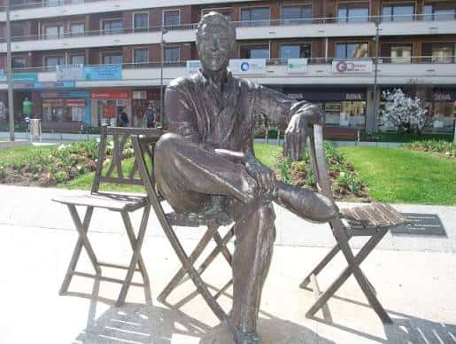 Estatua de bronce de Luis Mariano sentado en una silla de jardín frente a comercios locales