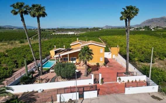 Vista aérea de una gran casa amarilla con piscina y palmeras en un entorno rural