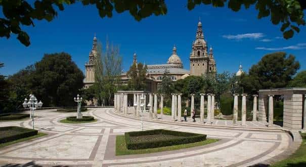 Plaza central de los jardines con senderos curvos, fuentes y la silueta de la catedral bajo un cielo azul