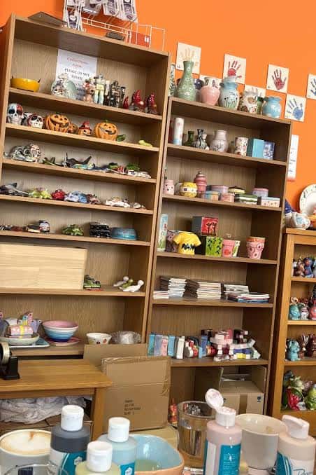 Interior del café mostrando estanterías con cerámicas pintadas y materiales para talleres