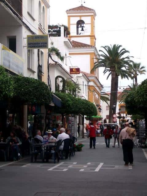 Calle peatonal con terrazas de cafeterías y la torre de una iglesia al fondo