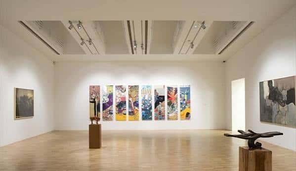 Sala de exposiciones con esculturas en pedestales y obra pictórica central multicolor