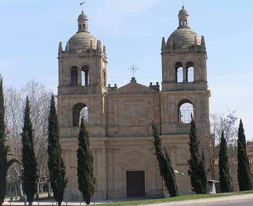 Fachada frontal de la Iglesia Nueva del Arrabal flanqueada por cipares altos y el cielo azul de fondo