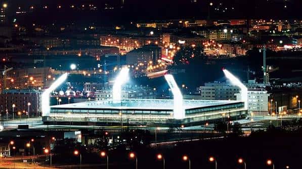 Estadio Nueva Balastera iluminado por la noche, destacando sus estructuras de iluminación sobre el campo y la ciudad al fondo