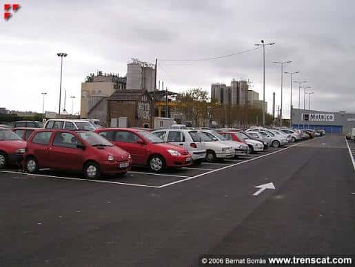 Vista general del aparcamiento de la estación con vehículos estacionados y edificios industriales al fondo