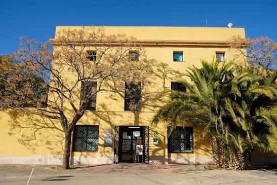 Vista frontal del Centre Cívic Mas Abelló con fachada amarilla y palmeras bajo cielo azul
