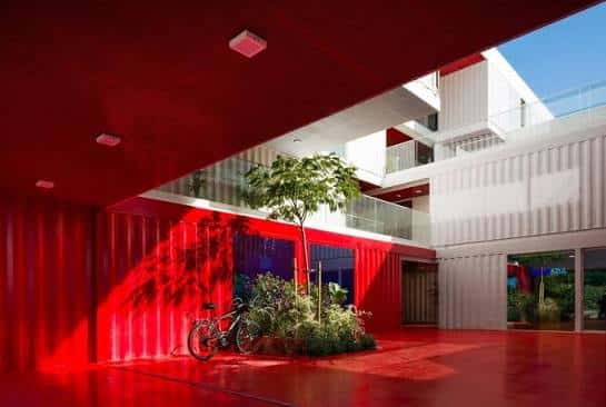 Área exterior cubierta con paredes rojas, vegetación y bicicleta frente a la fachada de contenedores