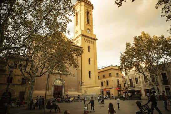 Plaza animada con árboles frondosos y la torre de la iglesia al atardecer, mostrando vida cotidiana en el espacio público